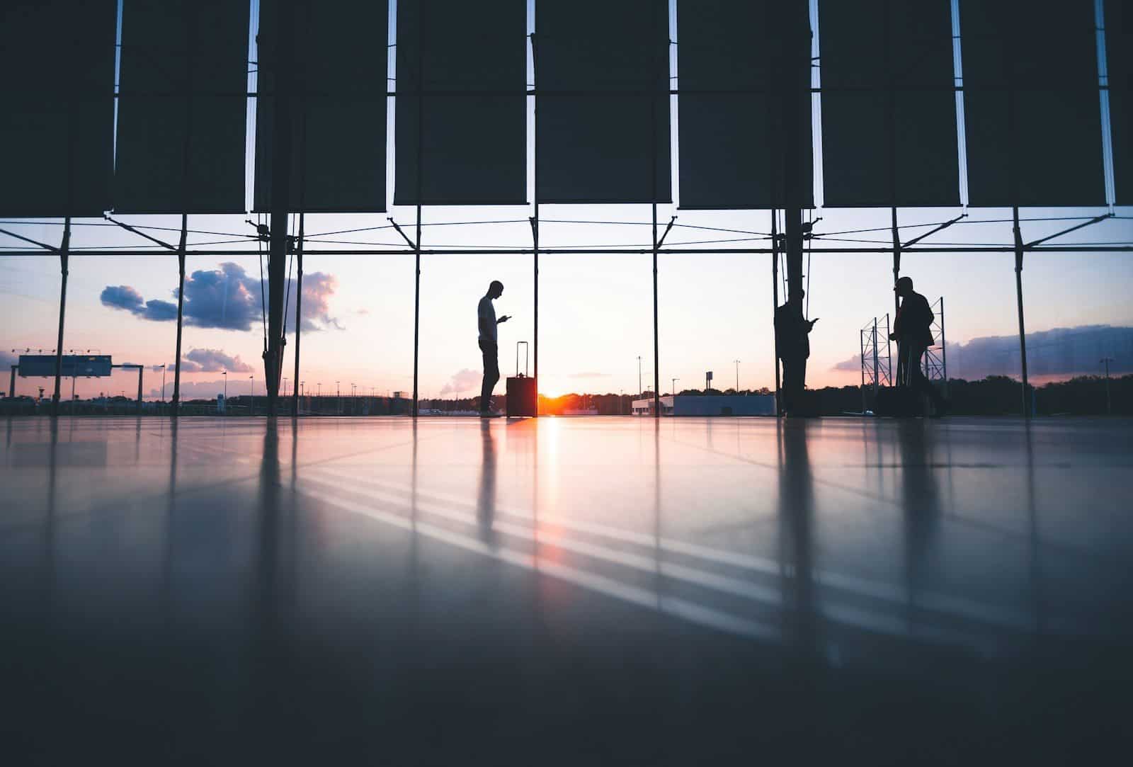 Silhouettes of people with luggage in a terminal against a bright low sun